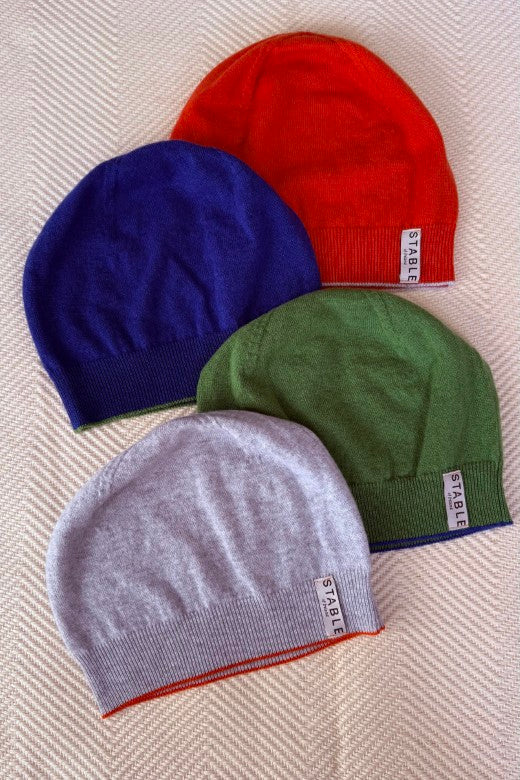 Four colorful STABLE Irish-made cashmere beanies featuring signature trim, displayed in orange, blue, green, and grey.