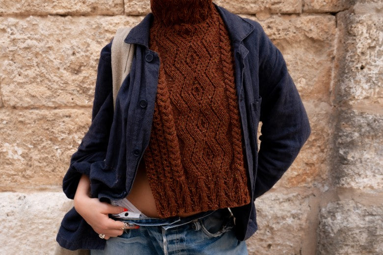 Hand-knit brown Aran cable bib by Irish maker Muriel, featuring intricate wool textures worn over a dark jacket.