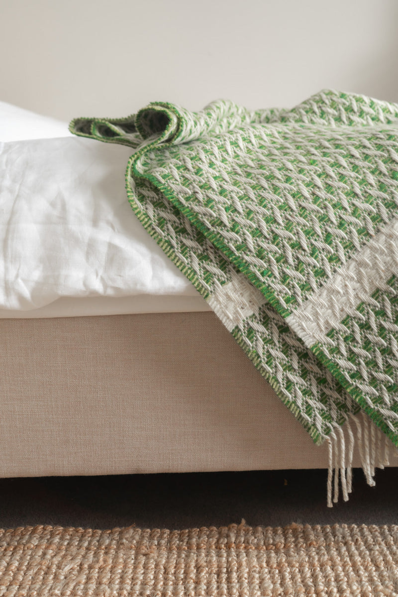 Light moss green Irish-made wicker blanket featuring a textured herringbone weave and fringe on a neutral bed.